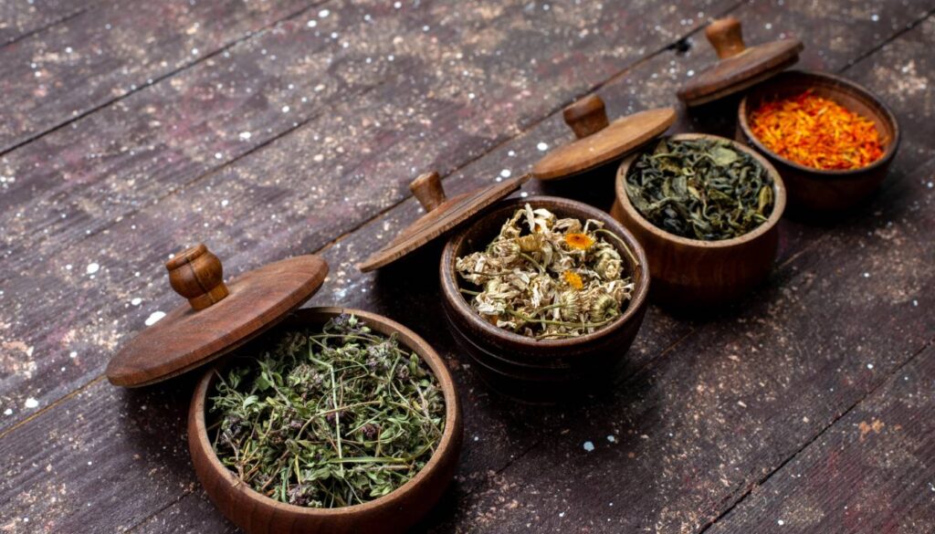 Four wooden bowls with lids, each containing different dried herbs, arranged in a row on a rustic wooden surface.