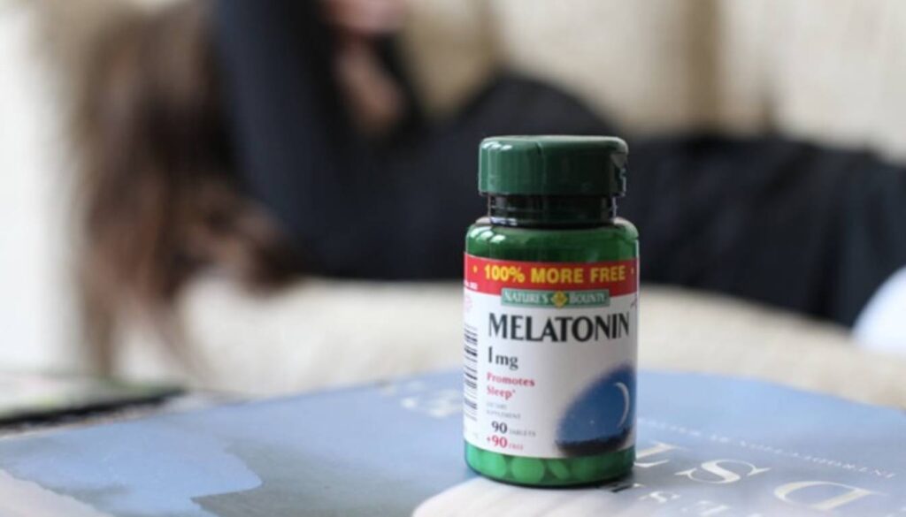 A bottle of Nature's Bounty Melatonin 1 mg tablets, labeled "Promotes Sleep," with a blurred background of a person lying on a couch.