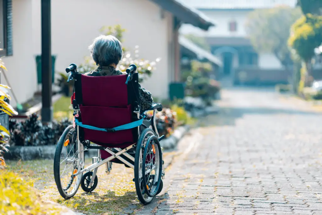 An elderly person in a wheelchair on a cobblestone street near a house with a garden.