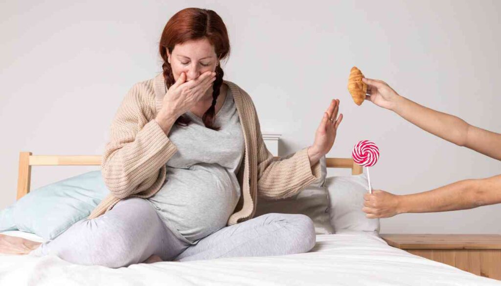 A pregnant woman sitting on a bed, holding her stomach with one hand and covering her mouth with the other, indicating nausea.