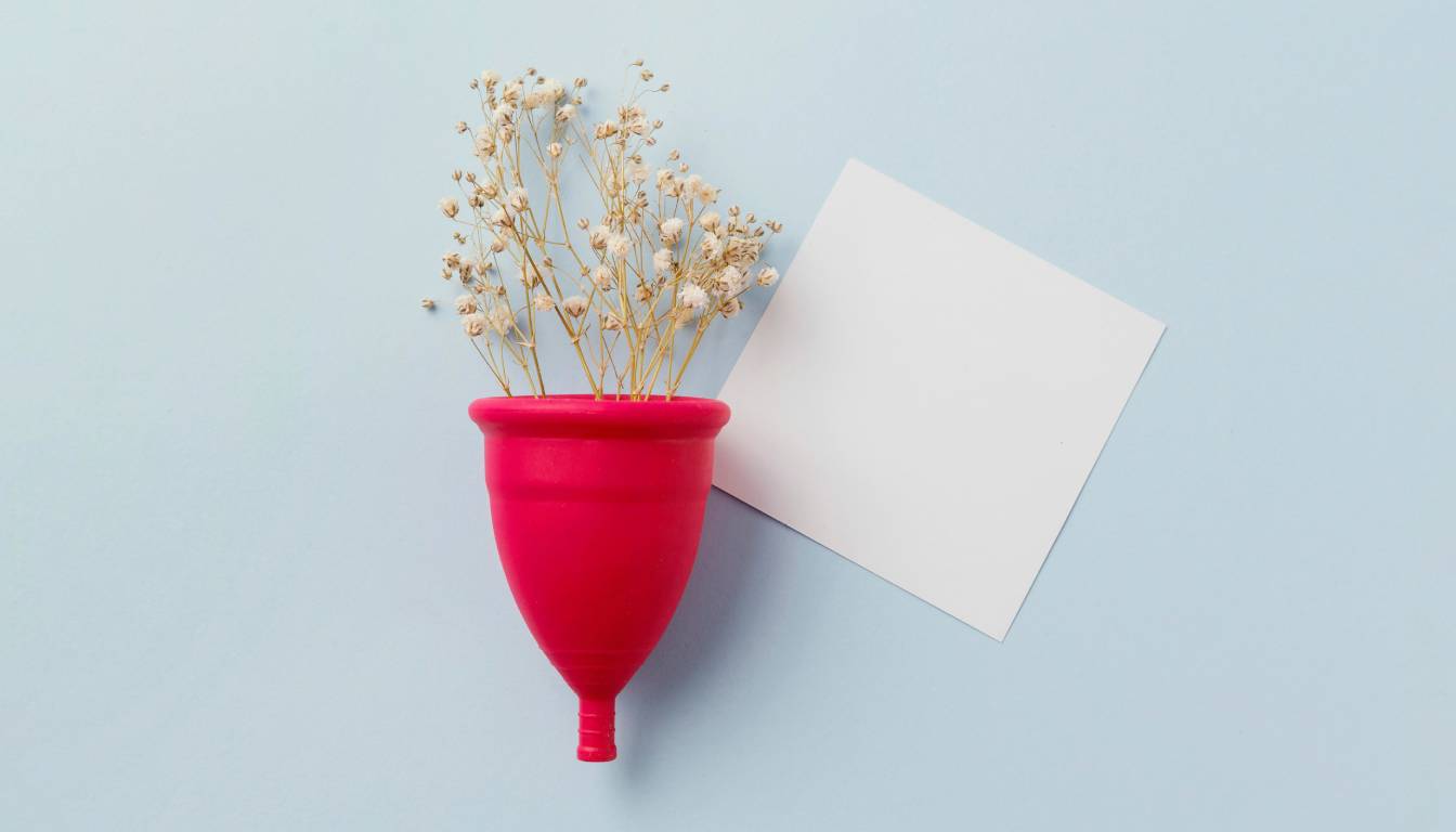 A red menstrual cup filled with small white flowers, placed on a light blue background next to a blank white card.