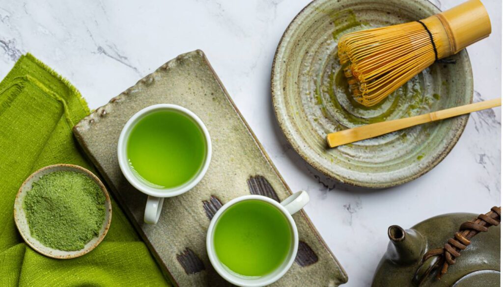 A traditional Japanese tea set with two cups of vibrant green matcha tea, a bamboo whisk, a bowl of matcha powder, and a teapot on a marble surface.