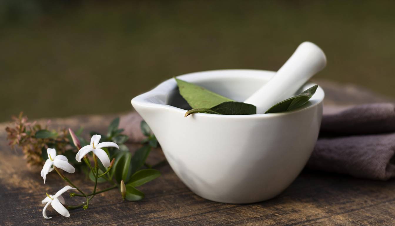 A white mortar and pestle with green leaves inside, accompanied by white flowers on a wooden surface.