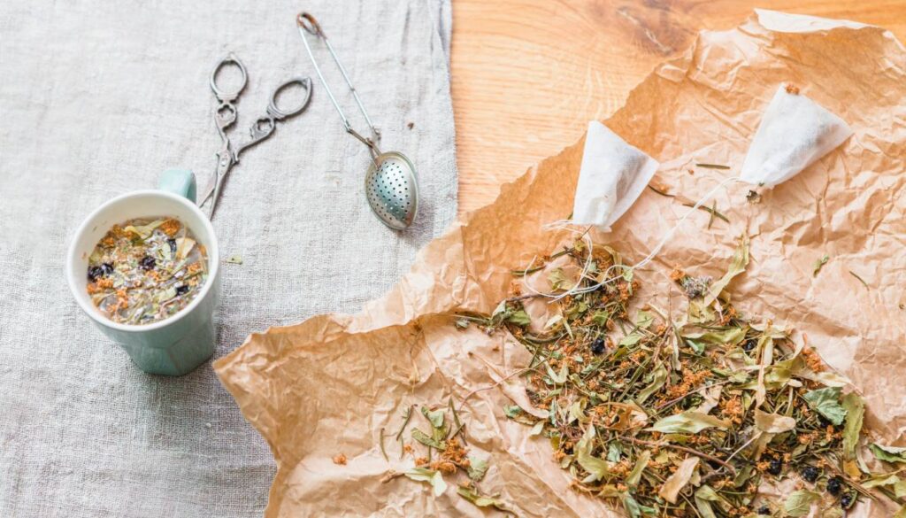 A cup of herbal tea, loose dried herbs, and tea-making tools on a linen cloth.