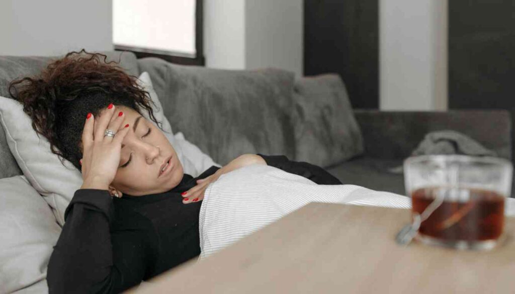 A person resting on a couch with their face obscured, holding their head, with a glass of dark liquid on the table nearby.