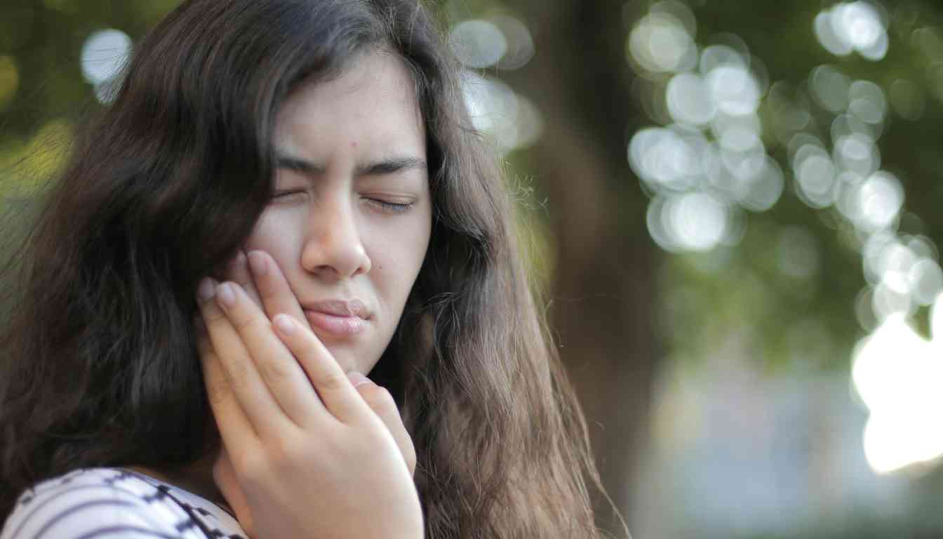 A person holding their jaw, possibly in pain, with a blurred background suggesting a natural outdoor setting.