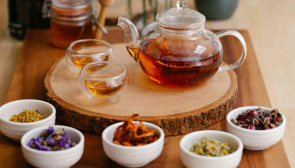 A transparent teapot filled with amber-colored tea sits on a wooden slice, surrounded by small white bowls containing various dried herbs and flowers, suggesting natural herbal remedies for the lymph system.