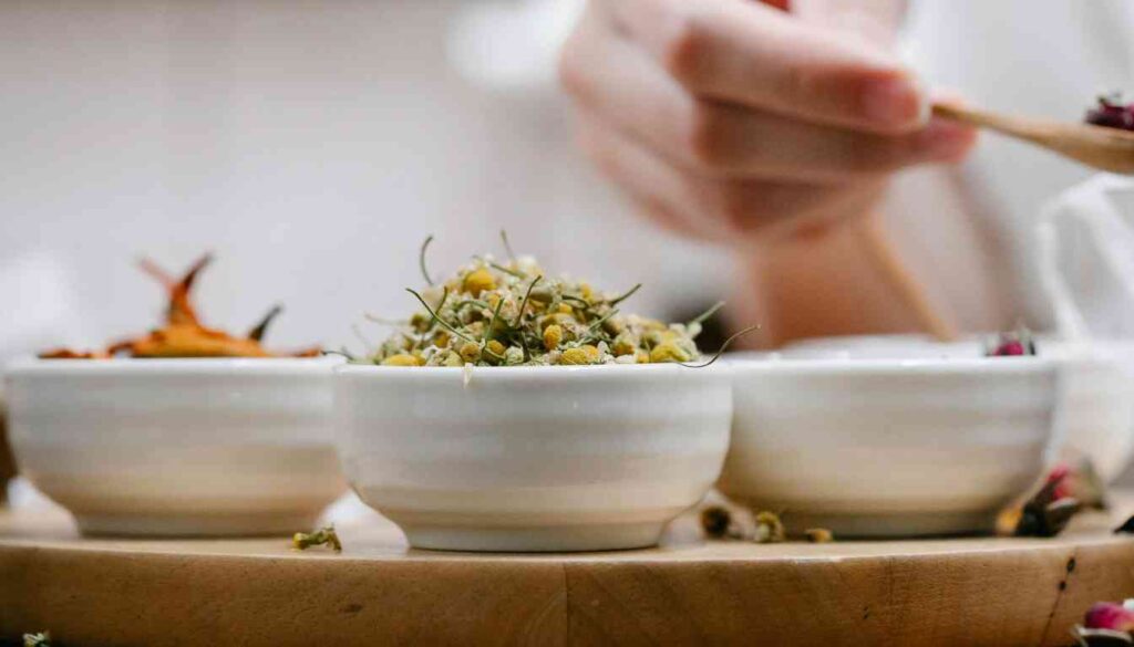 Three white bowls on a wooden surface, each containing different types of dried herbs. The bowl in the foreground is filled with small yellow buds, the middle bowl has larger orange pieces, and the rear bowl contains a mix of petals and leaves.