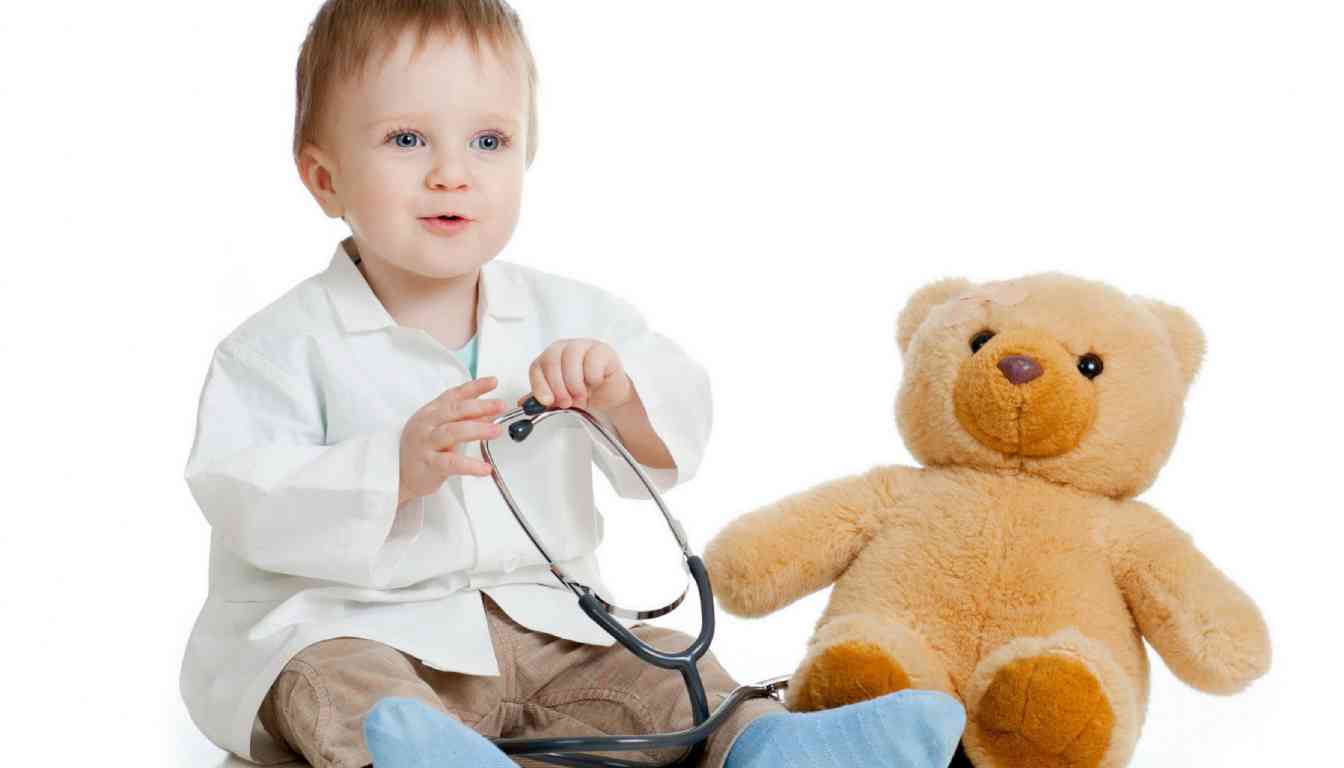 A young child dressed in a white coat holding a stethoscope, sitting next to a teddy bear.