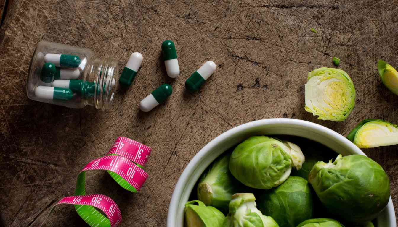 A wooden surface with a jar of green and white capsules, a pink measuring tape, and a bowl of Brussels sprouts.