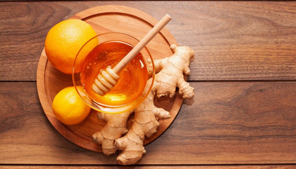 A wooden board with a bowl of honey, a honey dipper, fresh ginger roots, and two citrus fruits, one orange and one lemon.