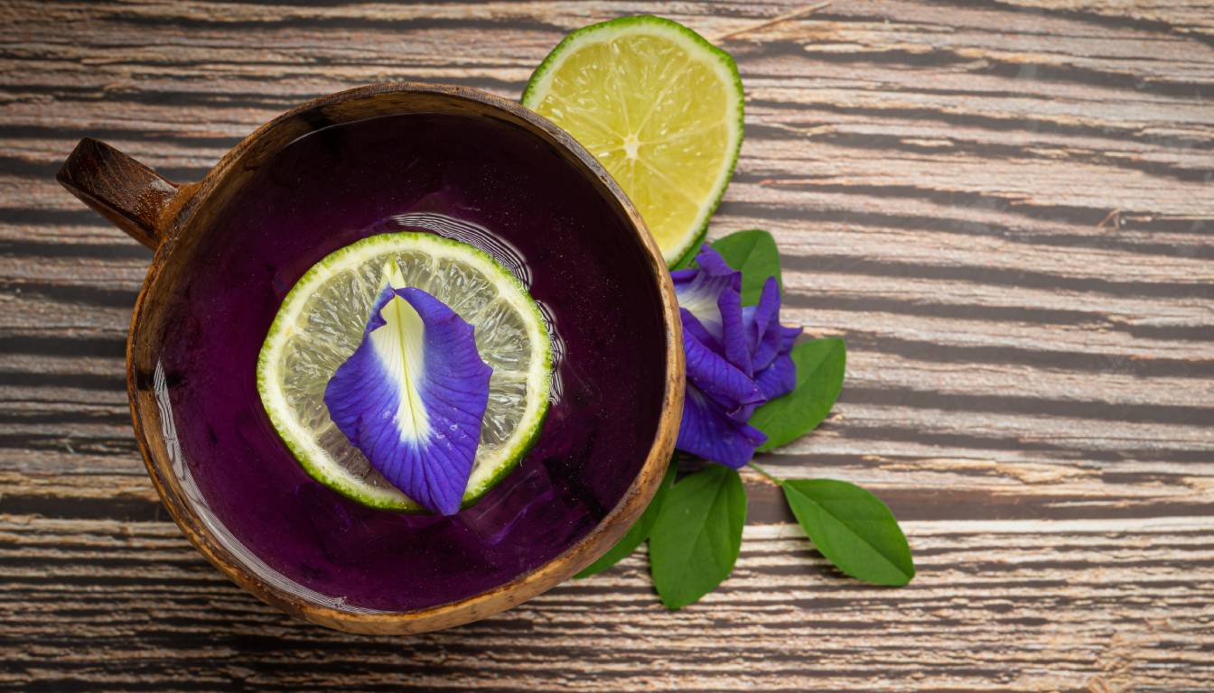 A wooden cup filled with a vibrant purple butterfly pea flower tea, garnished with a slice of lime and a butterfly pea flower petal. The cup is placed on a wooden surface with additional lime slices and butterfly pea flowers nearby.