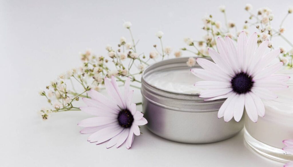 An open tin of healing salve surrounded by delicate white and purple flowers, including daisies and baby's breath, on a white background.