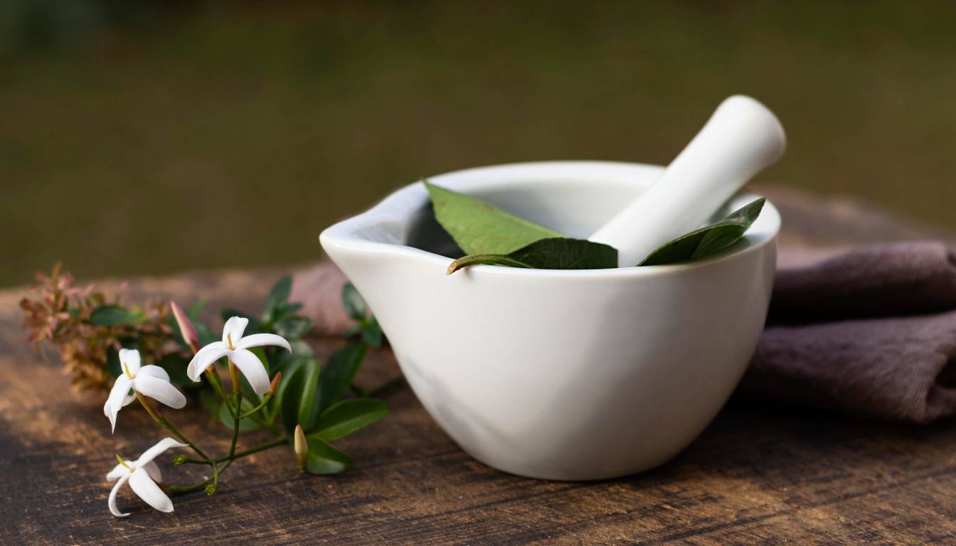 A white ceramic mortar and pestle with green leaves inside, placed on a wooden surface next to a small branch with white flowers.