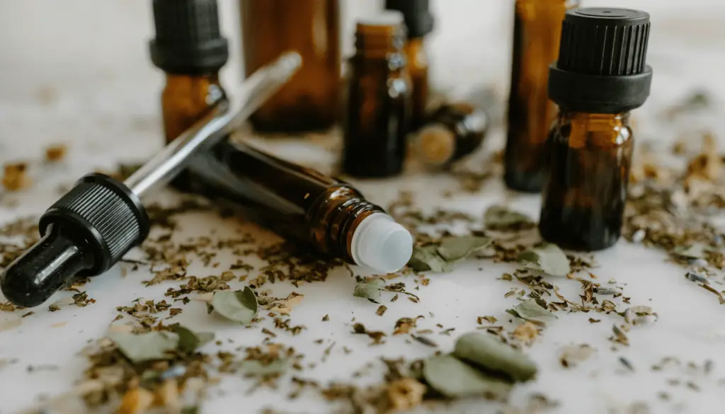 A collection of small brown glass bottles with droppers, some lying on their sides, surrounded by scattered dried herbs on a light surface.