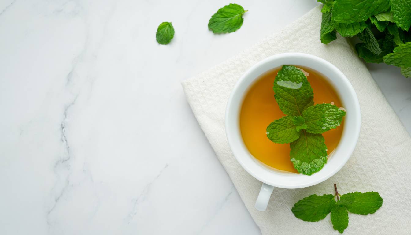 A white cup filled with herbal mint tea, garnished with fresh mint leaves, placed on a white cloth napkin with additional mint leaves scattered around.