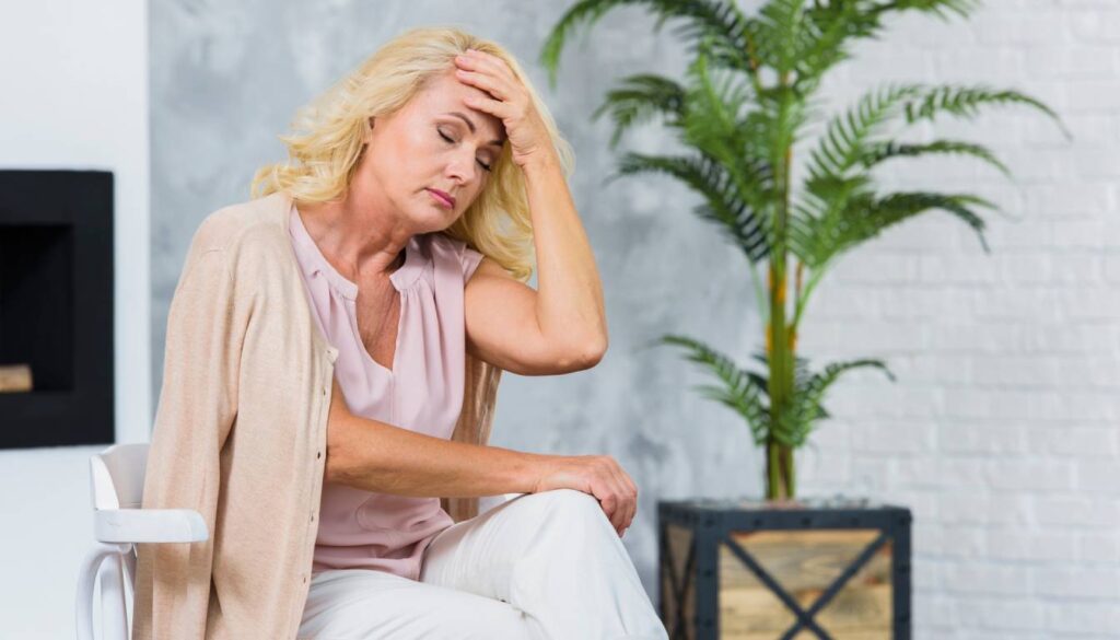 A woman sitting with her hand on her forehead, appearing contemplative or concerned, with a potted plant in the background.