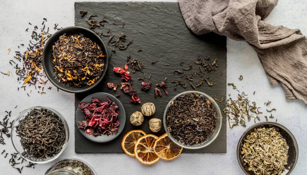 A variety of dried herbs and tea leaves are displayed in bowls on a dark slate surface, accompanied by dried citrus slices and a textured cloth.
