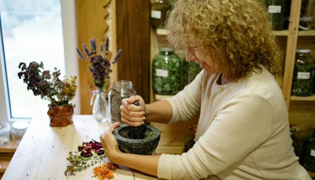 A person using a mortar and pestle to grind herbs on a wooden table, surrounded by jars of dried herbs and flowers.