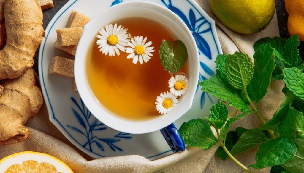 A cup of herbal tea garnished with chamomile flowers and mint leaves, surrounded by fresh ginger, sugar cubes, lemon, and mint sprigs.