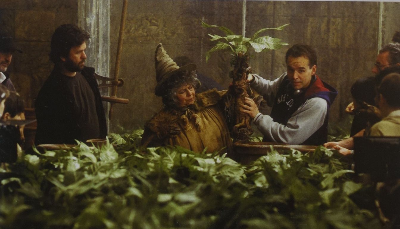 A group of people in a greenhouse setting, surrounded by lush green plants, with one person holding a large plant with a humanoid figure resembling a scarecrow.