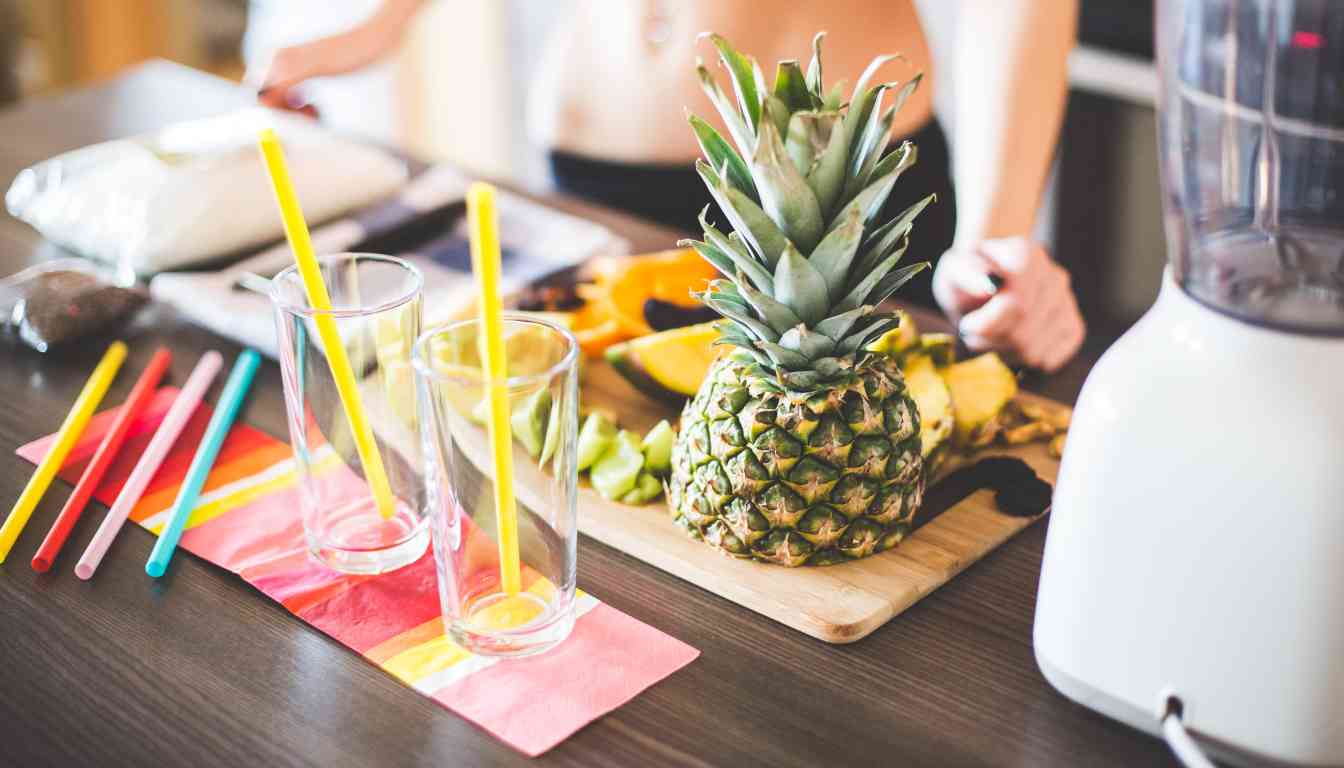 A kitchen counter with ingredients and utensils for making a Herbalife protein shake. Visible are a whole pineapple, bananas, avocados, colorful straws, an empty glass, and a blender.