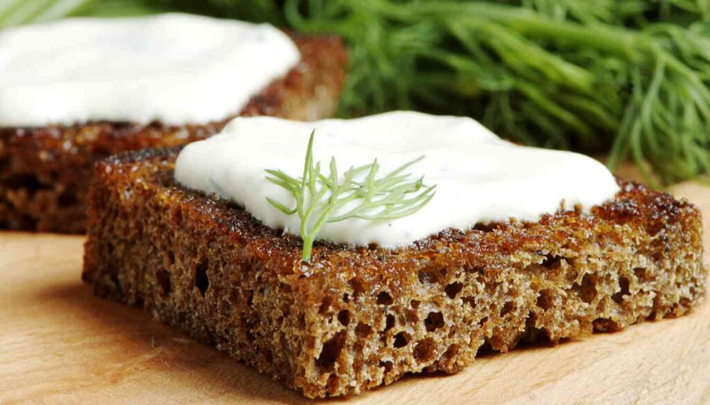 Two slices of brown bread topped with creamy white dill dressing and garnished with a sprig of fresh dill, with more dill in the background.