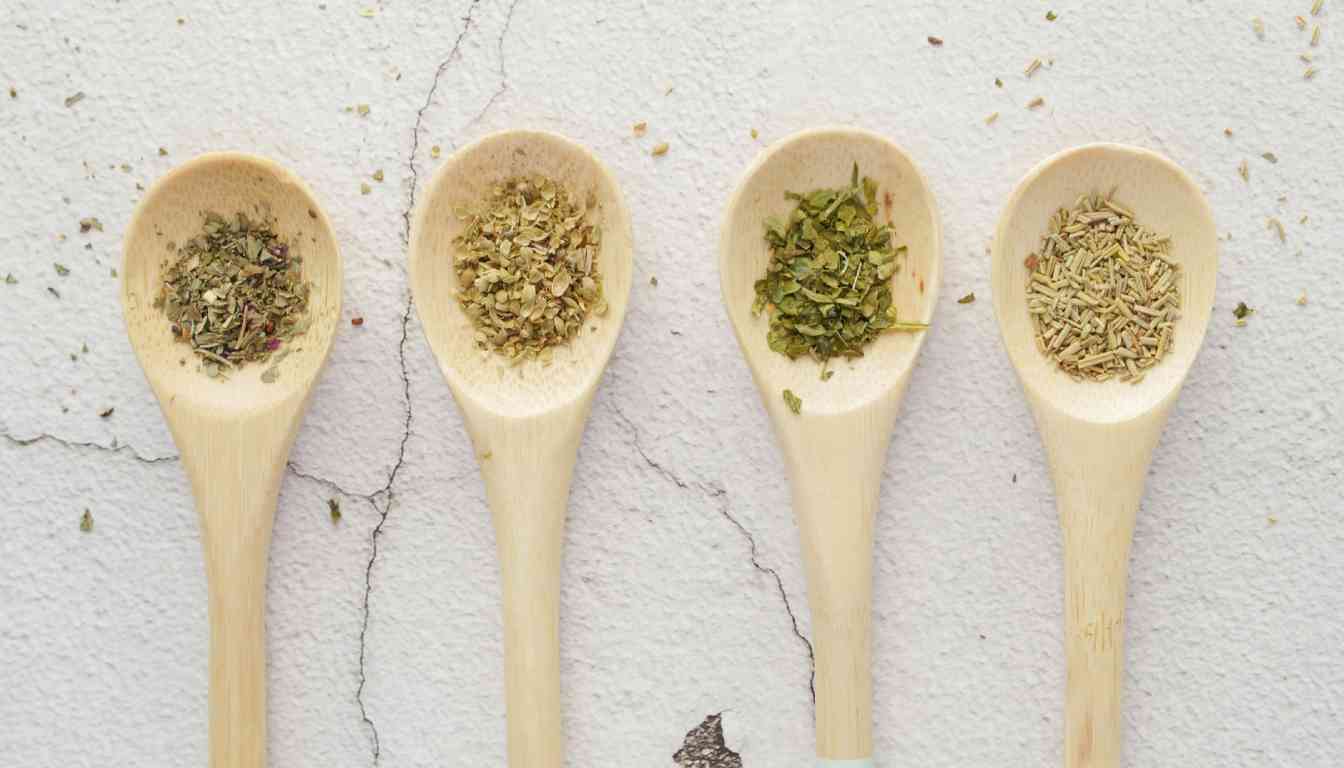 Four wooden spoons with different dried herbs on a light textured surface.