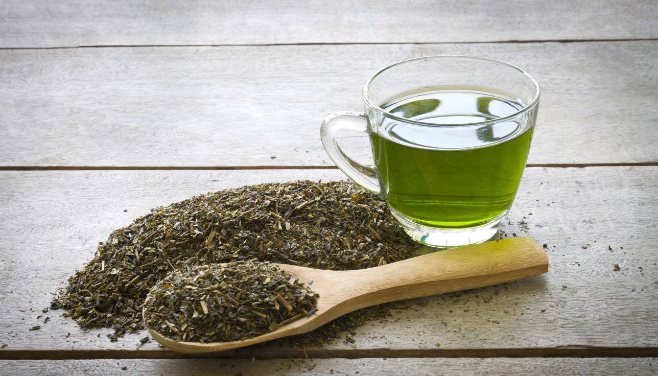 A glass cup filled with green tea sits on a wooden surface next to a pile of dried green tea leaves and a wooden spoon.