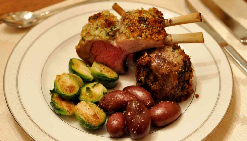 A succulent herb-crusted rack of lamb served with roasted brussels sprouts and boiled red-skinned potatoes on a white plate.