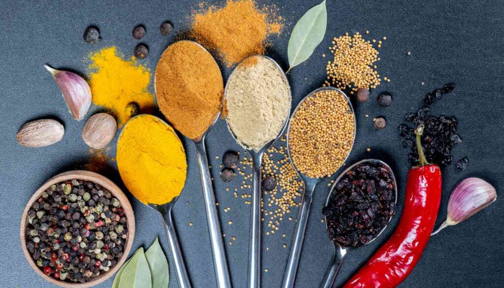 An array of spices known for their anti-inflammatory properties displayed on a dark surface. The collection includes turmeric, ginger, cinnamon, black peppercorns, cloves, mustard seeds, and chili peppers.