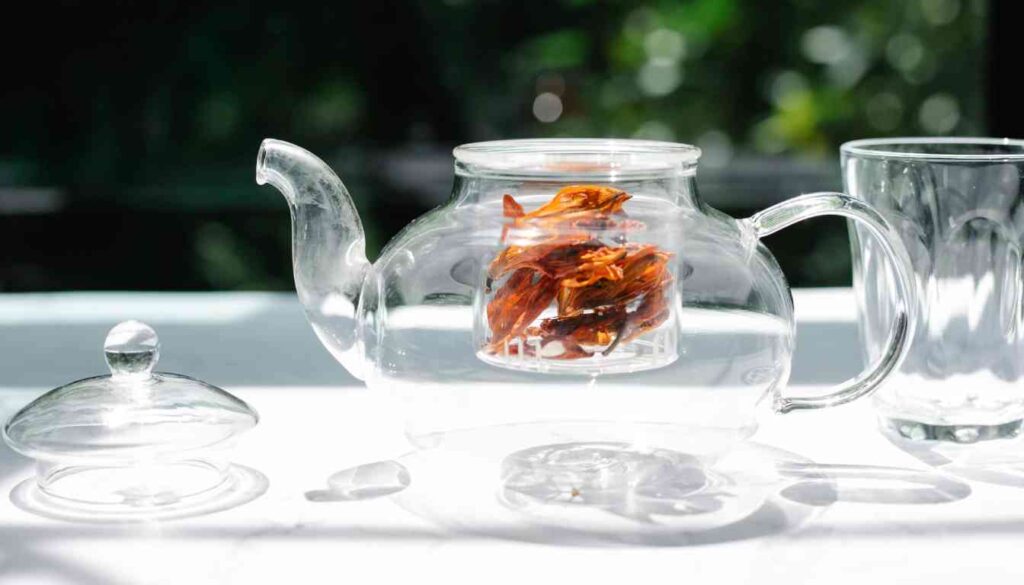 A clear glass teapot with vibrant orange herbal leaves steeping inside, accompanied by a matching clear glass cup, all set against a blurred green background suggesting an outdoor setting.