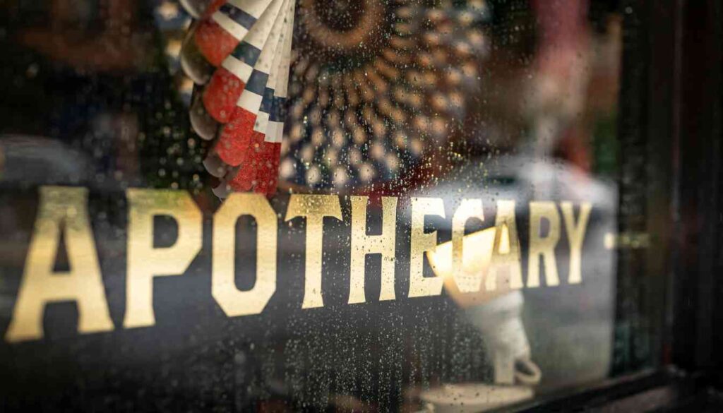 Close-up of a vintage glass window with the word “APOTHECARY” painted in gold lettering, reflecting an American flag, suggesting a traditional or historical pharmacy setting.