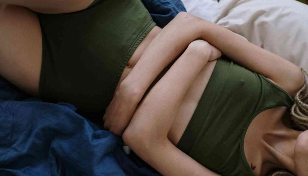 A person lying on a bed, wearing green underwear and a matching green top, with hands placed on the lower abdomen, suggesting they might be experiencing menstrual cramps.