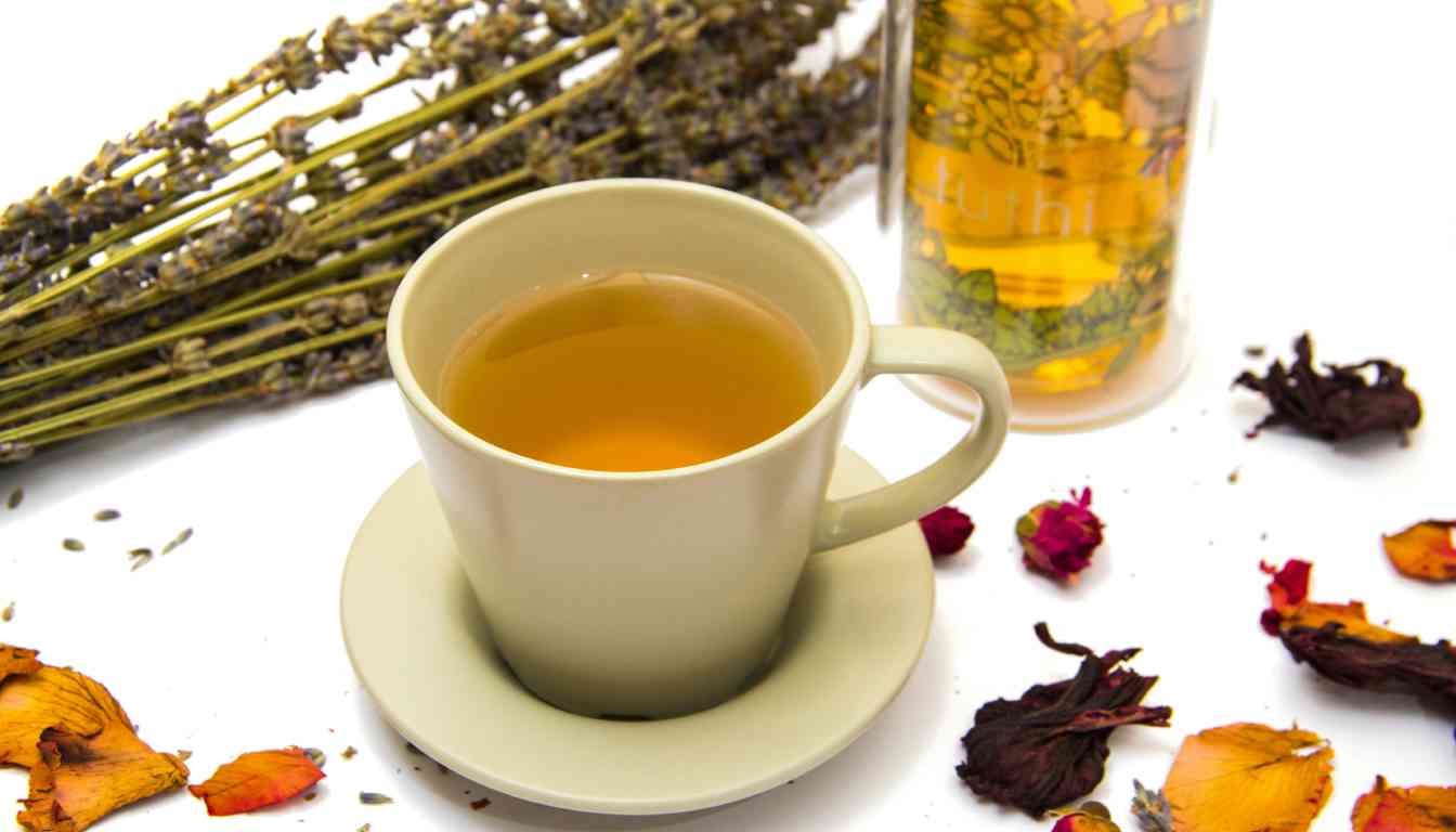 A cup of herbal tea on a saucer surrounded by dried herbs and petals, with a bottle of dried lavender stalks in the background on a white surface.