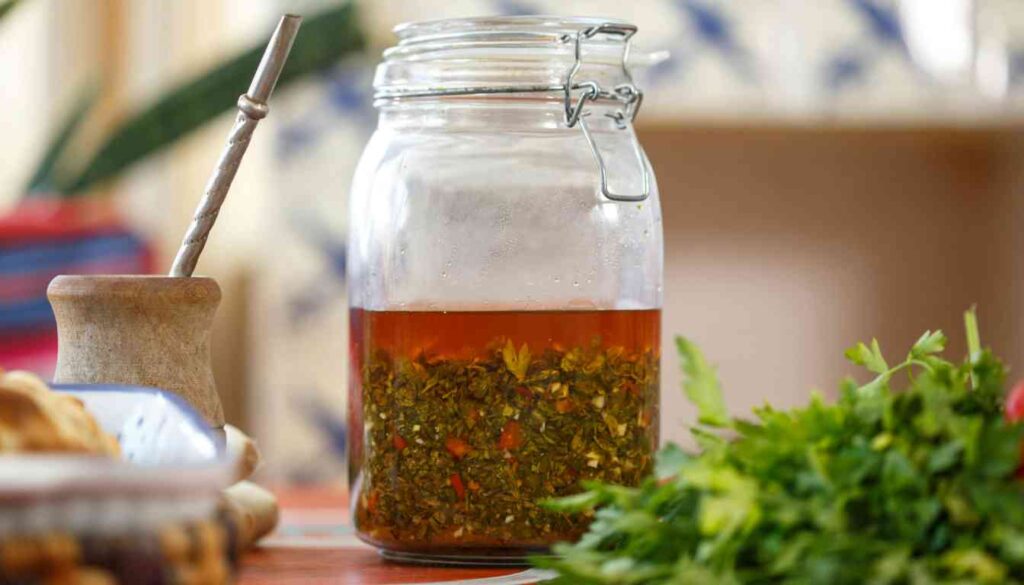 A glass jar filled with herb and garlic marinade, with a wooden mortar and pestle and fresh herbs in the foreground.