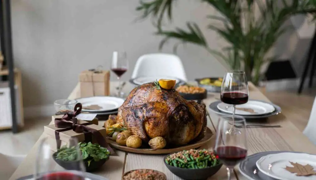 A golden-brown herb-roasted turkey on a wooden platter garnished with herbs and lemon, surrounded by side dishes, with two glasses of red wine on a dining table.