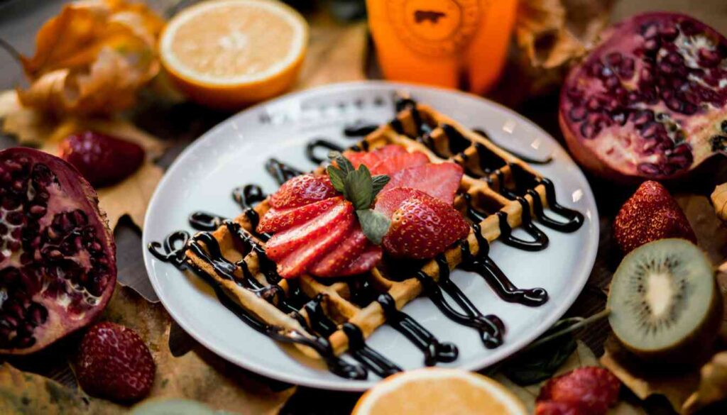 A plate of freshly made waffles topped with sliced strawberries and drizzled with chocolate syrup, surrounded by an assortment of fruits including pomegranate, kiwi, and oranges, suggesting a healthy and appetizing Herbalife waffle recipe.