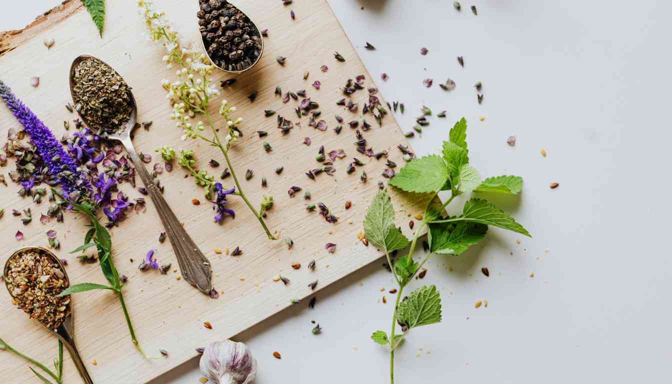 A variety of herbs and spices are scattered on a wooden surface with a focus on a wooden cutting board. On the board, there are several spoons filled with different seasonings, including what appears to be dried lavender, black peppercorns, and possibly a mixed herb seasoning. Fresh green leaves and purple flowers add a pop of color to the composition.