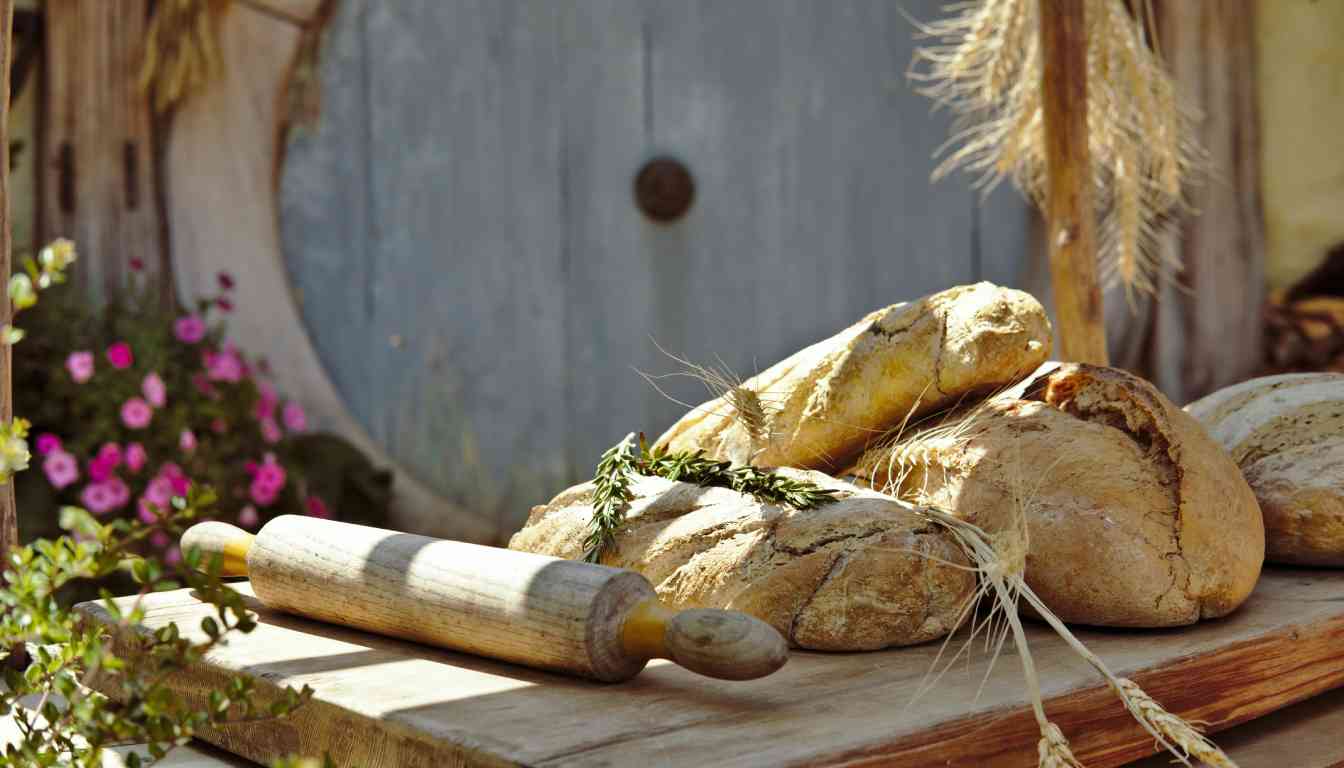 Freshly baked herbal bread loaves on a wooden board with a rolling pin, wheat stalks, and green herbs in the foreground, set against a rustic background with a wooden door and blooming flowers.