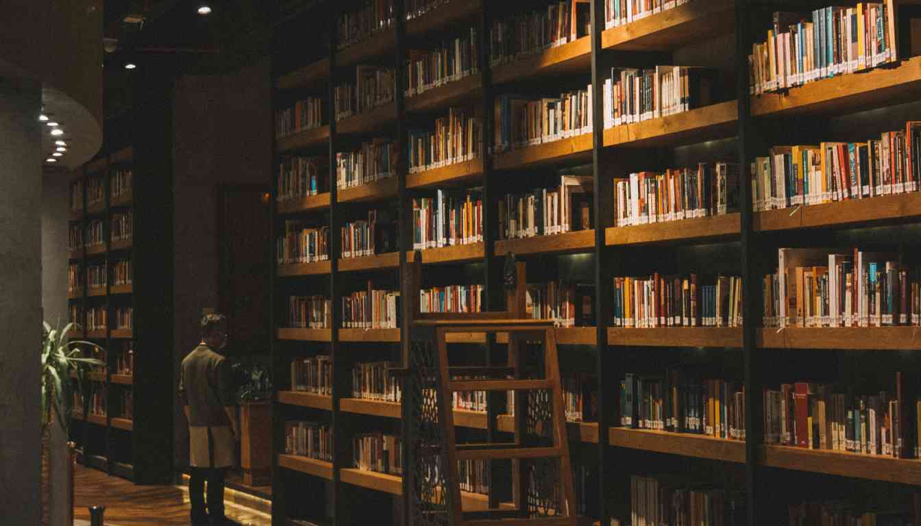 A person stands in a dimly lit library with tall bookshelves filled with books. The library has a warm, cozy atmosphere with wooden shelves and soft lighting.