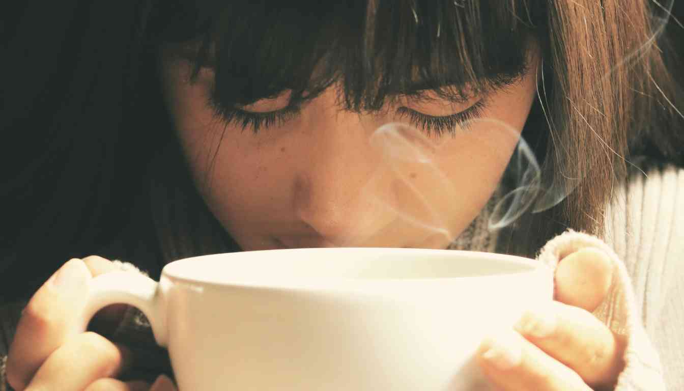 Close-up of an individual with closed eyes inhaling steam from a large white mug, possibly as a home remedy for sinus relief.