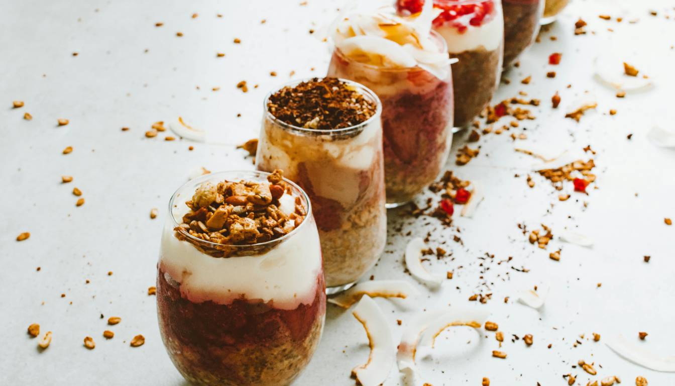 A row of parfait glasses filled with layered ingredients such as granola, yogurt, and fruit, arranged on a white surface with scattered granola and coconut flakes.