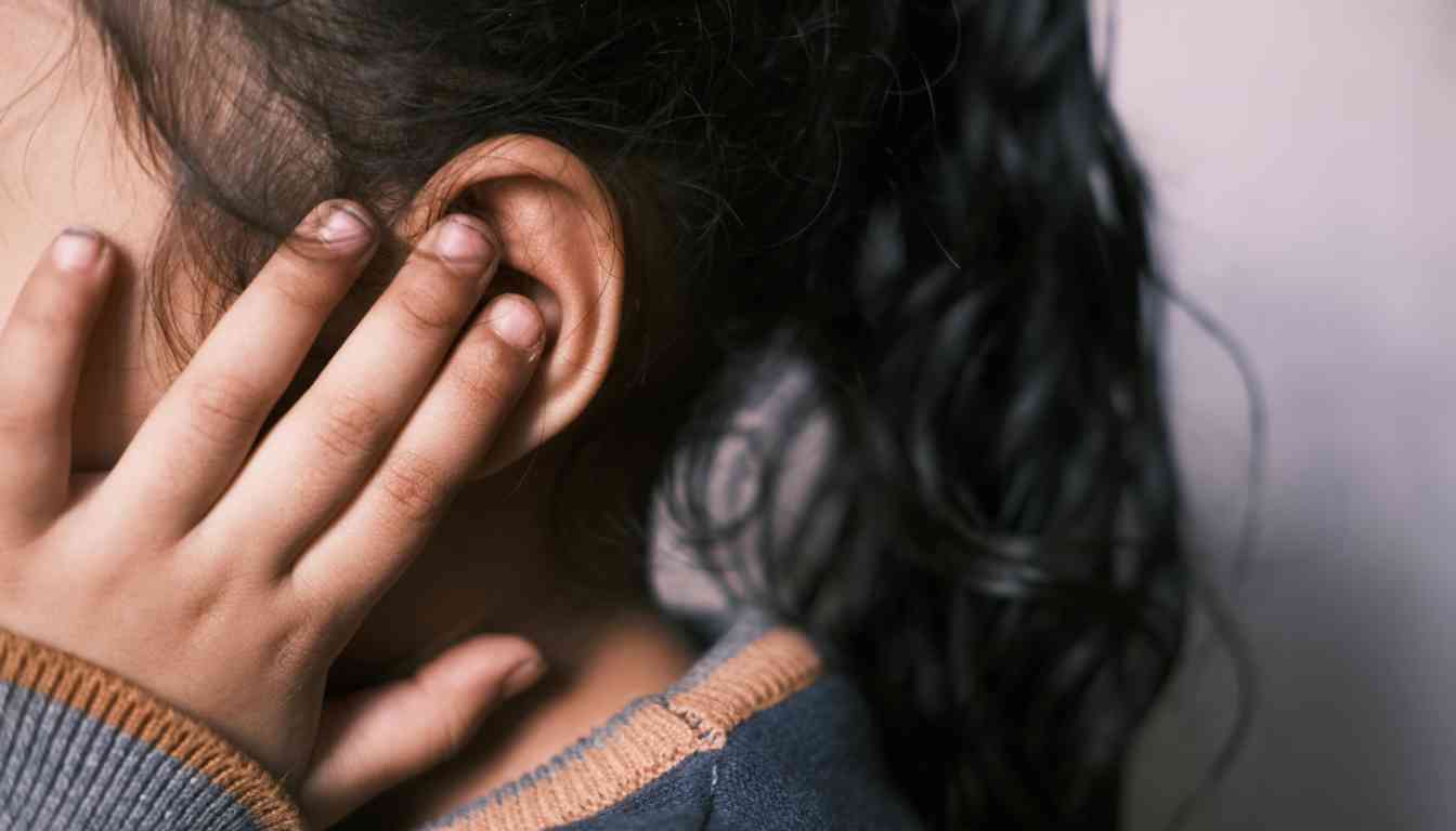 A person holding their left ear, possibly indicating discomfort or pain, with a focus on the ear and hand.