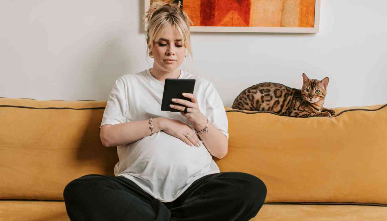 A pregnant person sitting on a yellow couch, holding a tablet, with a Bengal cat lying beside them.