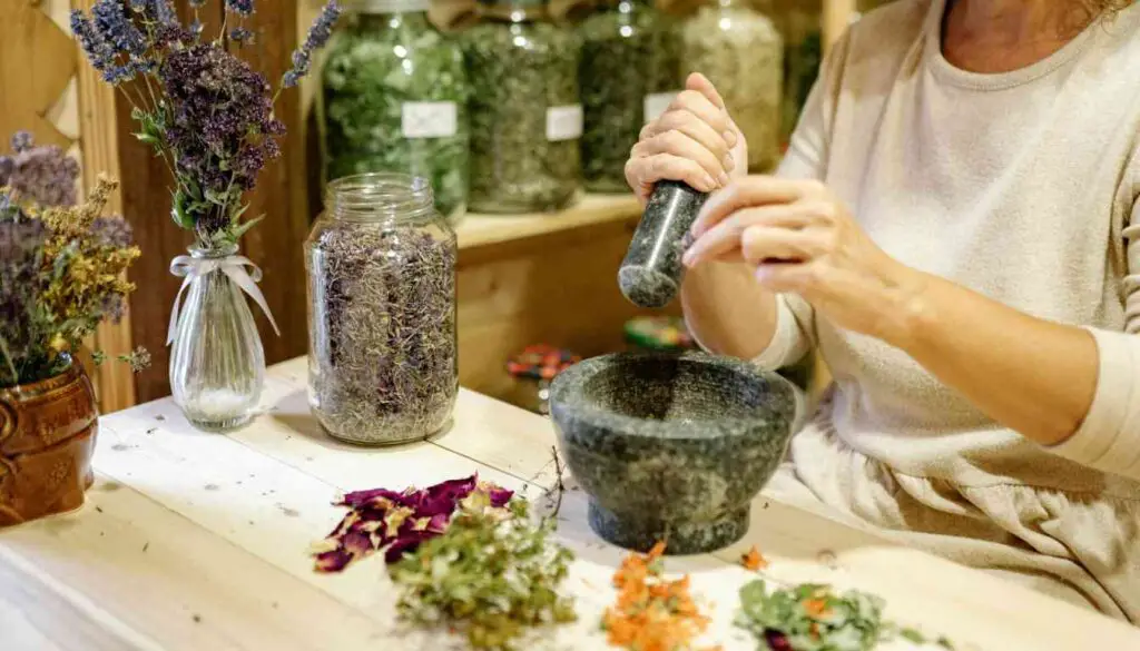 A person preparing herbal medicine using a mortar and pestle, surrounded by jars of dried herbs and fresh plants on a wooden table.