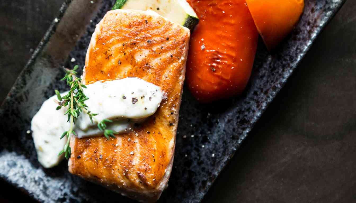 A succulent piece of herb-crusted salmon served with a dollop of creamy sauce, garnished with fresh thyme on a black rectangular plate, accompanied by grilled vegetables.