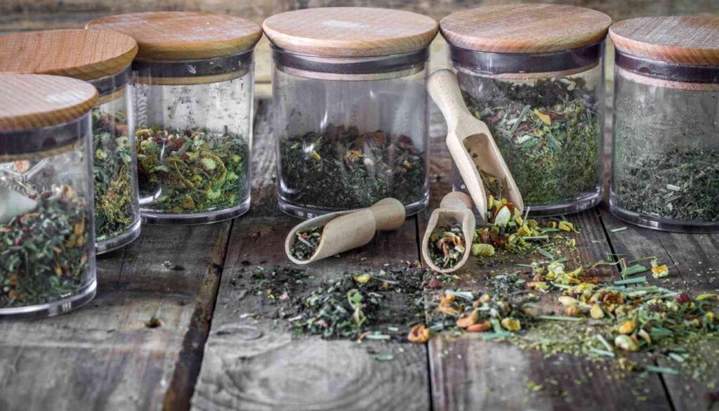 Several glass jars with wooden lids containing dried herbs, with some herbs spilled on a rustic wooden surface and two wooden scoops filled with herbs.