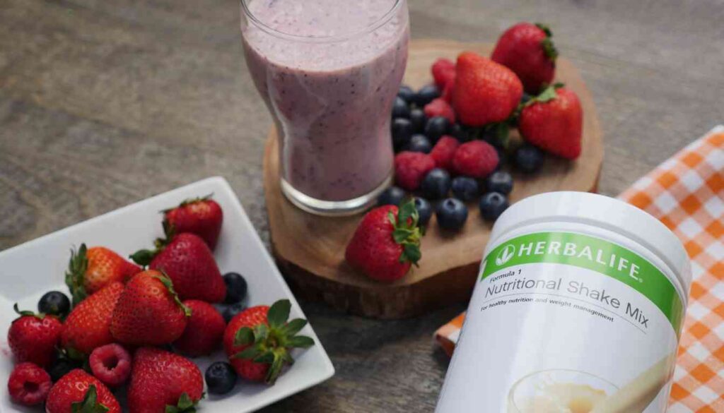 A glass of pink smoothie on a wooden stand surrounded by fresh strawberries, raspberries, and blueberries. Next to it is an open Herbalife Nutritional Shake Mix container.