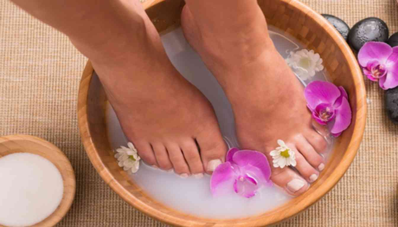 A pair of feet soaking in a wooden bowl of water with floating flowers, suggesting a foot soak treatment possibly for tinea pedis.